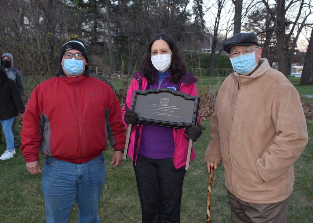 Two men and one woman stading outside, wearing masks. Woman in center is holding a palacque